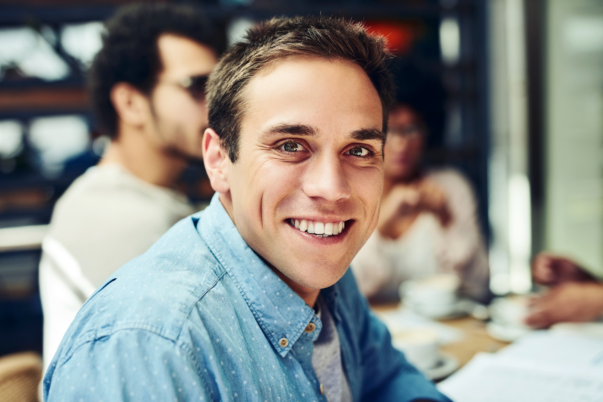 A young man with short brown hair and a blue button-up shirt smiles warmly at the camera. He is seated in a casual setting, possibly a café, with blurred individuals and tables in the background creating a cozy, social atmosphere.