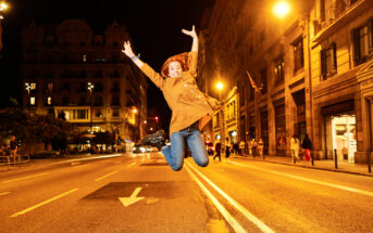 A person jumps joyfully in the middle of a brightly lit street at night, surrounded by tall buildings and streetlights. Their arms are raised high, and they wear a brown jacket and blue jeans. The street is empty except for a few pedestrians in the background.