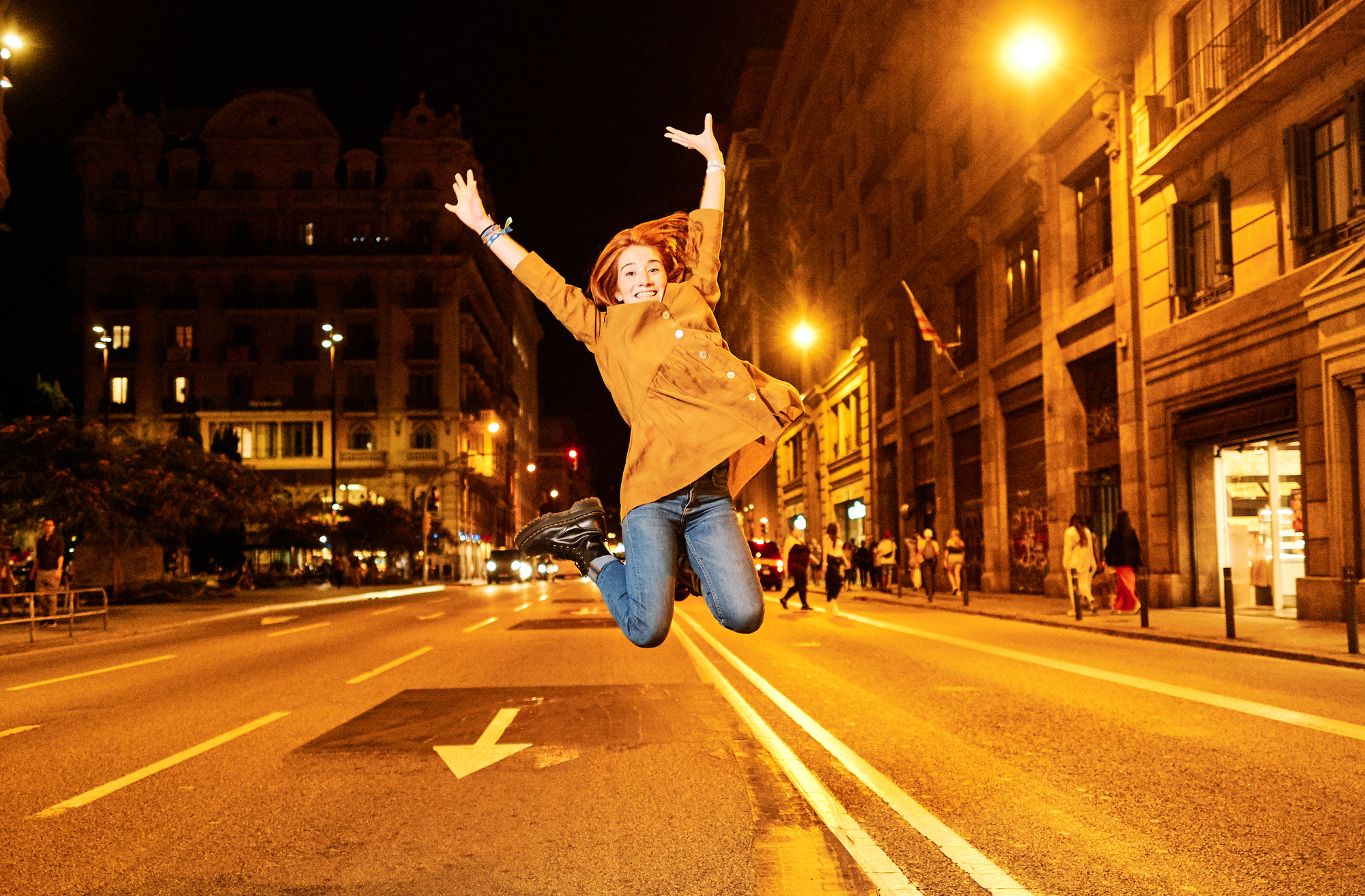 A person jumps joyfully in the middle of a brightly lit street at night, surrounded by tall buildings and streetlights. Their arms are raised high, and they wear a brown jacket and blue jeans. The street is empty except for a few pedestrians in the background.