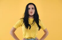 A person with long dark hair stands against a bright yellow background. They are wearing a yellow short-sleeved top and have their hands on their hips. Their expression is serious.