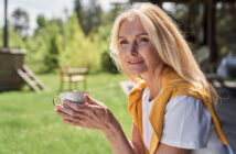 A woman with long blond hair holds a cup while sitting outdoors on a sunny day. She wears a white shirt with a yellow sweater draped over her shoulders and smiles gently at the camera. Trees and a green lawn are visible in the background.