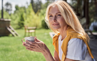 A woman with long blond hair holds a cup while sitting outdoors on a sunny day. She wears a white shirt with a yellow sweater draped over her shoulders and smiles gently at the camera. Trees and a green lawn are visible in the background.