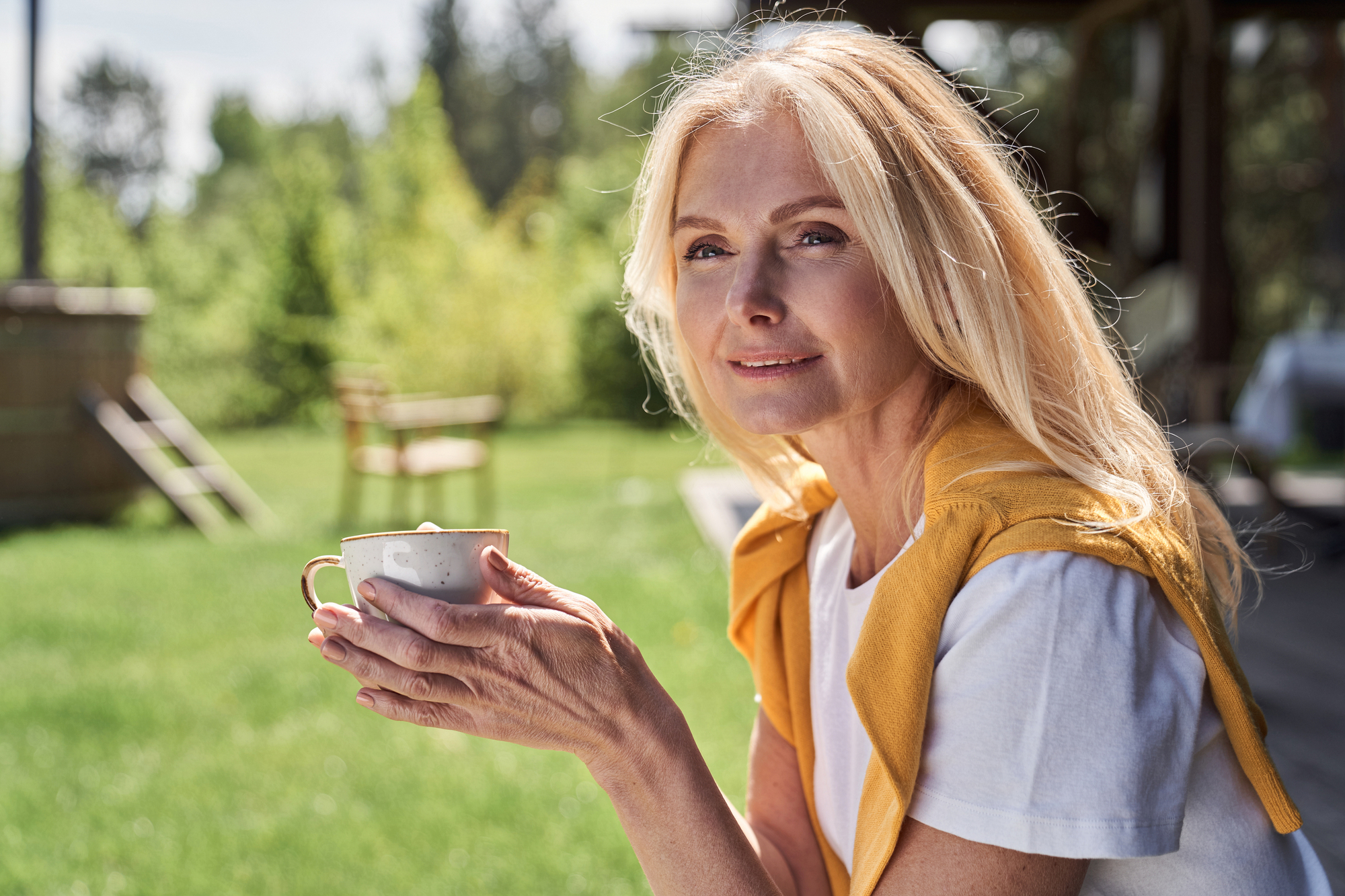 A woman with long blond hair holds a cup while sitting outdoors on a sunny day. She wears a white shirt with a yellow sweater draped over her shoulders and smiles gently at the camera. Trees and a green lawn are visible in the background.