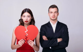 A woman in a red dress holds a broken red heart symbol with a sad expression, while a man in a black suit stands beside her with arms crossed, looking serious or indifferent. The background is a plain light gray.