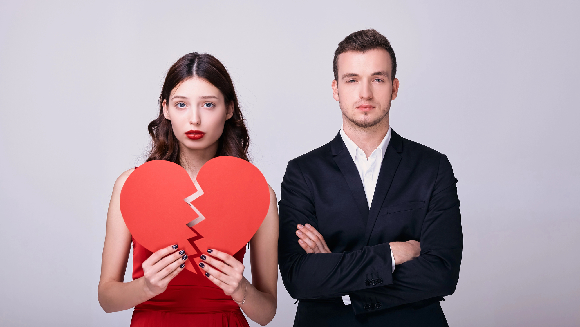 A woman in a red dress holds a broken red heart symbol with a sad expression, while a man in a black suit stands beside her with arms crossed, looking serious or indifferent. The background is a plain light gray.