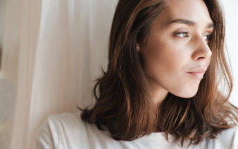 A woman with shoulder-length brown hair gazes thoughtfully out of a window. She is wearing a white shirt and stands in natural light, creating a serene and contemplative mood.
