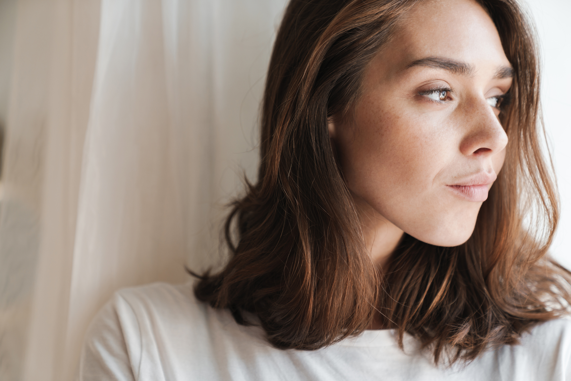 A woman with shoulder-length brown hair gazes thoughtfully out of a window. She is wearing a white shirt and stands in natural light, creating a serene and contemplative mood.