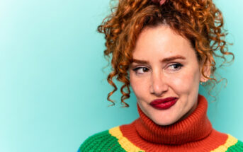 A woman with curly red hair styled in a ponytail is looking to her side thoughtfully. She is wearing a colorful sweater with a high orange turtleneck and bright red lipstick, standing against a light teal background.