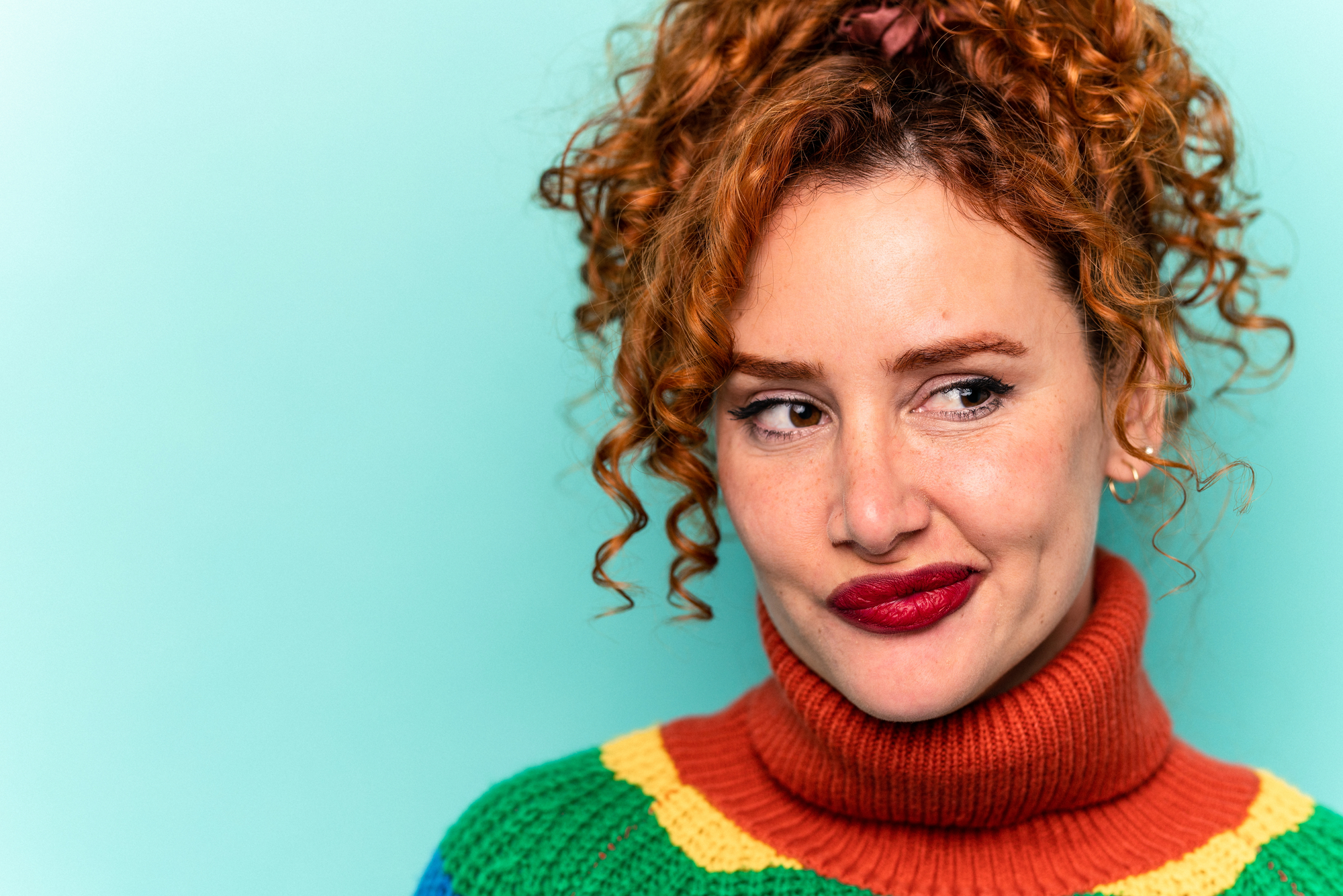 A woman with curly red hair styled in a ponytail is looking to her side thoughtfully. She is wearing a colorful sweater with a high orange turtleneck and bright red lipstick, standing against a light teal background.