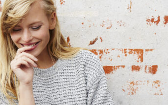 A blonde woman wearing a gray knitted sweater leans against a white brick wall with some red paint marks. She smiles and looks down, touching her lips gently with her fingers. Her hair is parted to the side, and she appears to be in a relaxed and cheerful mood.