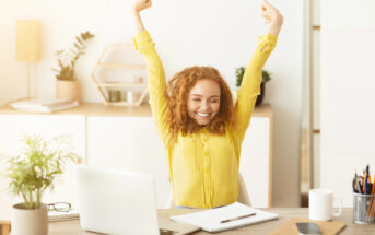 A woman with curly red hair, wearing a yellow shirt, sits at a desk with a laptop and notebooks. She has her arms raised in a victory pose, smiling with her eyes closed. The desk is adorned with plants and office supplies, and a shelf with decor is in the background.