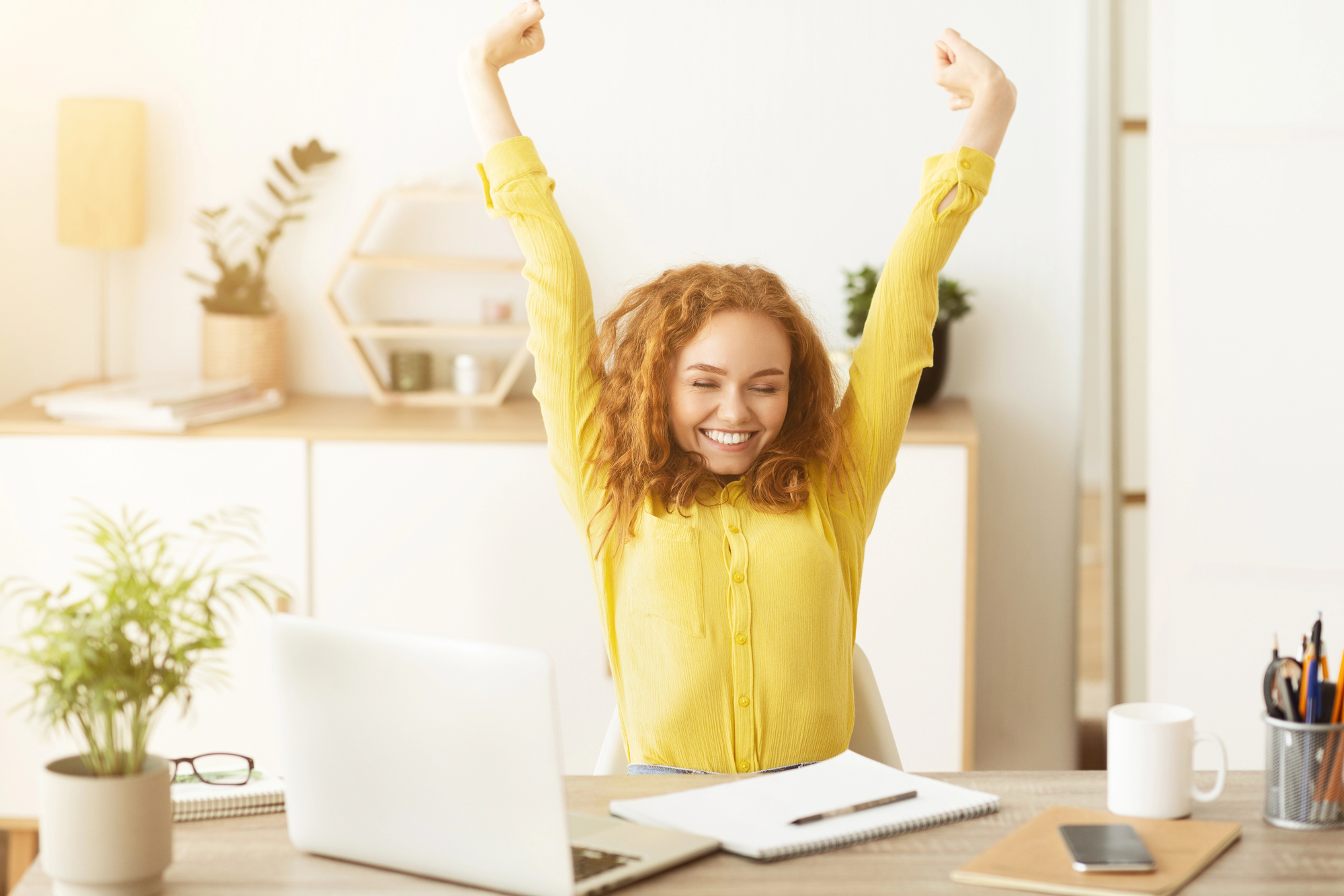 A woman with curly red hair, wearing a yellow shirt, sits at a desk with a laptop and notebooks. She has her arms raised in a victory pose, smiling with her eyes closed. The desk is adorned with plants and office supplies, and a shelf with decor is in the background.