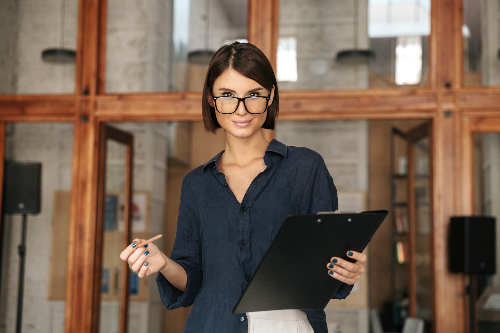 A woman with short, dark hair is standing indoors, holding a clipboard and a pen. She is wearing a dark blouse and glasses, looking confidently at the camera. The background features wooden-framed glass windows and shelves.