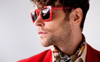 A person with curly hair and a well-groomed beard wears stylish red sunglasses and a red jacket. They are also adorned with a patterned scarf. The individual is looking confidently to the side against a neutral background.