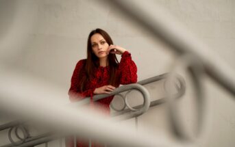 A woman with long brown hair wearing a red dress with black patterns leans casually on a gray railing. She is looking thoughtfully into the distance. The photo is framed with a soft focus on part of the railing in the foreground.