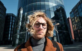 A person with wavy blond hair and round glasses is standing in front of a modern glass building, wearing a brown coat and gray turtleneck. The wind is blowing their hair, and the background includes other contemporary buildings against a clear blue sky.