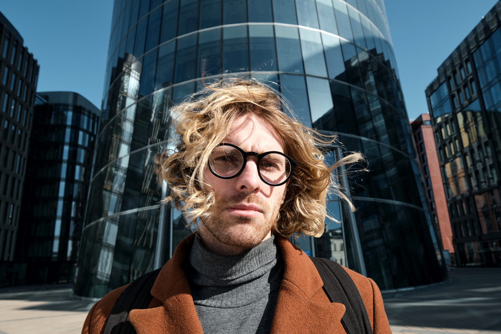 A person with wavy blond hair and round glasses is standing in front of a modern glass building, wearing a brown coat and gray turtleneck. The wind is blowing their hair, and the background includes other contemporary buildings against a clear blue sky.