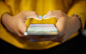 A close-up of a person wearing a yellow top, holding a smartphone with both hands and typing with their thumbs. They have light blue nail polish on their nails, and a beaded bracelet on one wrist is partially visible. The phone screen is glowing.