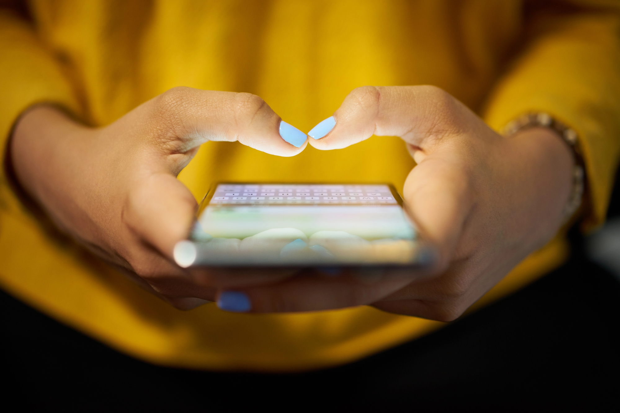 A close-up of a person wearing a yellow top, holding a smartphone with both hands and typing with their thumbs. They have light blue nail polish on their nails, and a beaded bracelet on one wrist is partially visible. The phone screen is glowing.