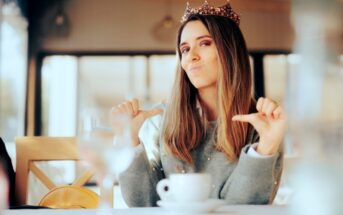 A woman with long hair, wearing a crown, sits at a table with a coffee cup in front of her. She is making a playful, confident expression while pointing at herself with both thumbs. The background is slightly blurred, suggesting an indoor café setting.