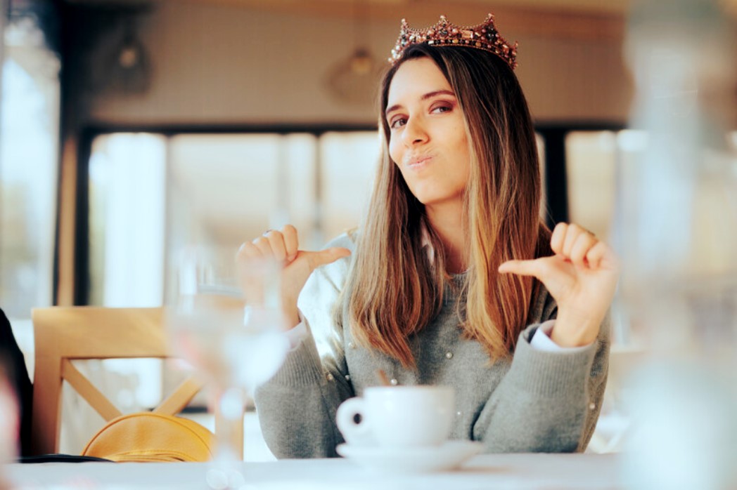 A woman with long hair, wearing a crown, sits at a table with a coffee cup in front of her. She is making a playful, confident expression while pointing at herself with both thumbs. The background is slightly blurred, suggesting an indoor café setting.