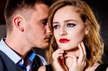 A man and a woman are close to each other in a dramatic scene. The man is whispering or speaking into the woman's ear while she looks forward with a concerned expression. The woman has red lipstick, winged eyeliner, and is holding her hands near her face.