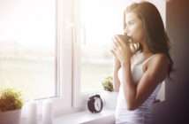A woman stands by a sunlit window, holding a mug with both hands and looking outside. She is dressed in a white tank top. On the windowsill, there is a small black alarm clock, two white candles, and a small plant.