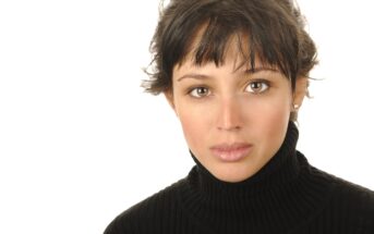 A woman with short, dark hair and bangs is wearing a black turtleneck sweater. She is looking directly at the camera with a neutral expression. The background is plain white. She also has small stud earrings.