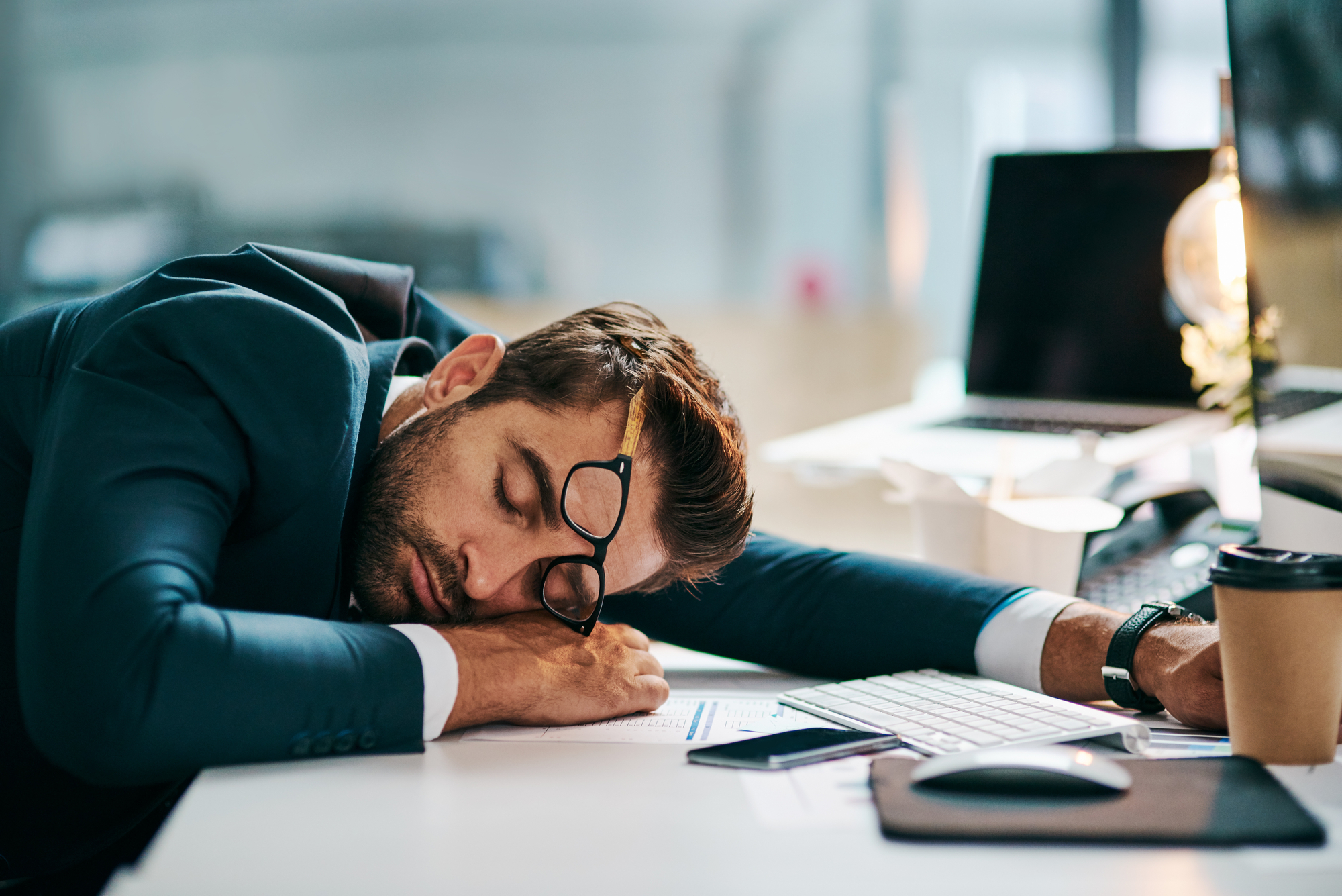 A man in a suit is sleeping at his desk with his head resting on papers. His glasses are askew, and his computer, smartphone, and a coffee cup are nearby, indicating he was working before falling asleep. The office setting appears modern and well-lit.