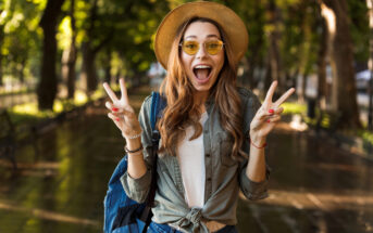 A young woman with long hair wearing a hat and yellow-tinted sunglasses smiles excitedly while holding up peace signs with both hands. She is dressed casually with a shirt and jeans and has a backpack on. The background shows a tree-lined path.