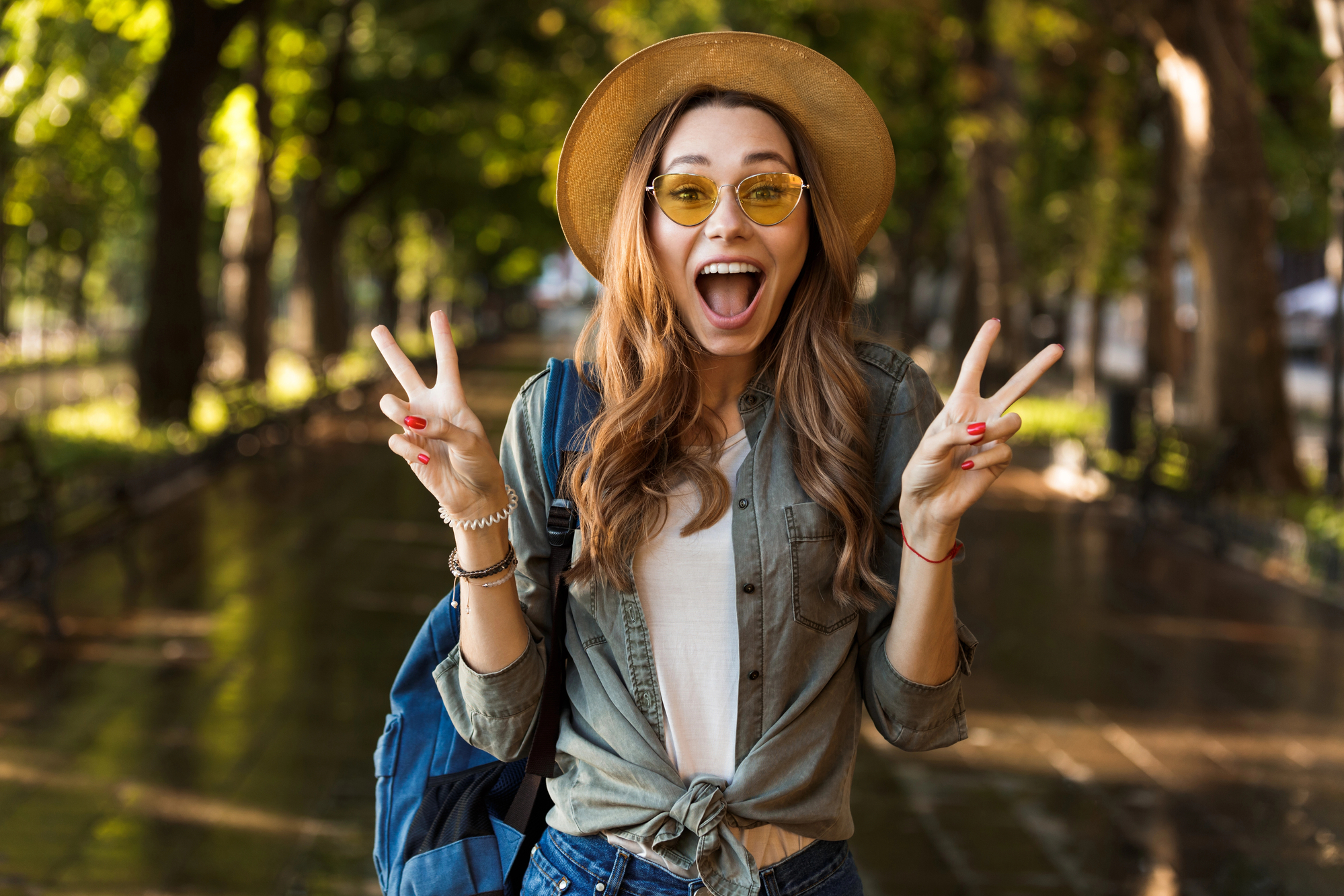 A young woman with long hair wearing a hat and yellow-tinted sunglasses smiles excitedly while holding up peace signs with both hands. She is dressed casually with a shirt and jeans and has a backpack on. The background shows a tree-lined path.