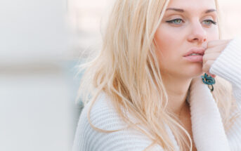 A young woman with long blonde hair is seen in a close-up shot. She is wearing a cozy white sweater and appears pensive, resting her chin on her hand. The background is blurred, emphasizing her thoughtful expression.