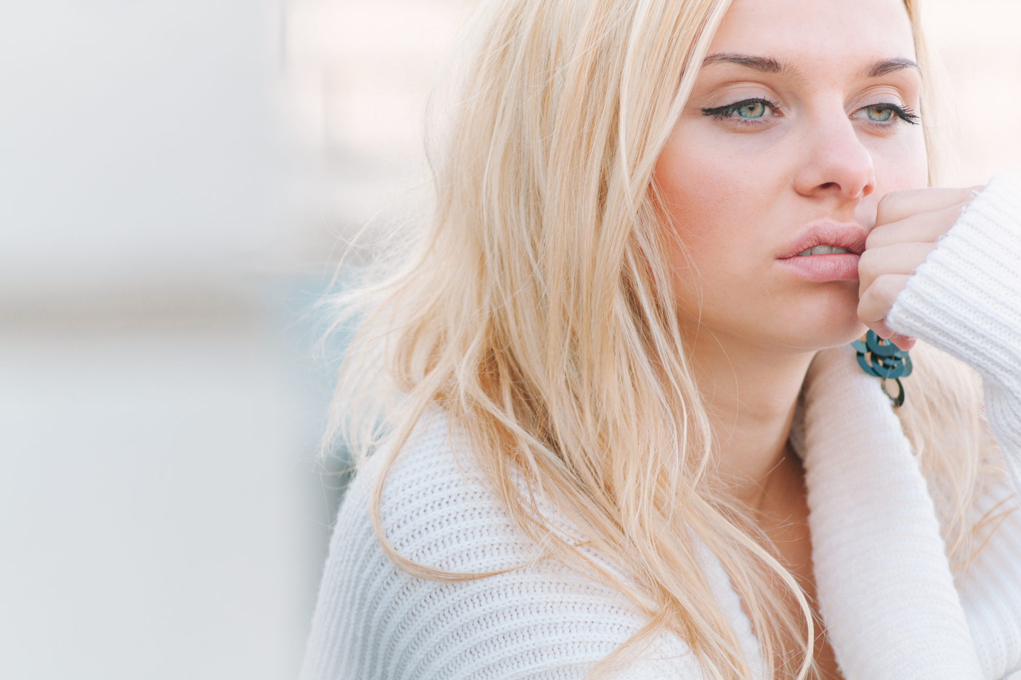 A young woman with long blonde hair is seen in a close-up shot. She is wearing a cozy white sweater and appears pensive, resting her chin on her hand. The background is blurred, emphasizing her thoughtful expression.