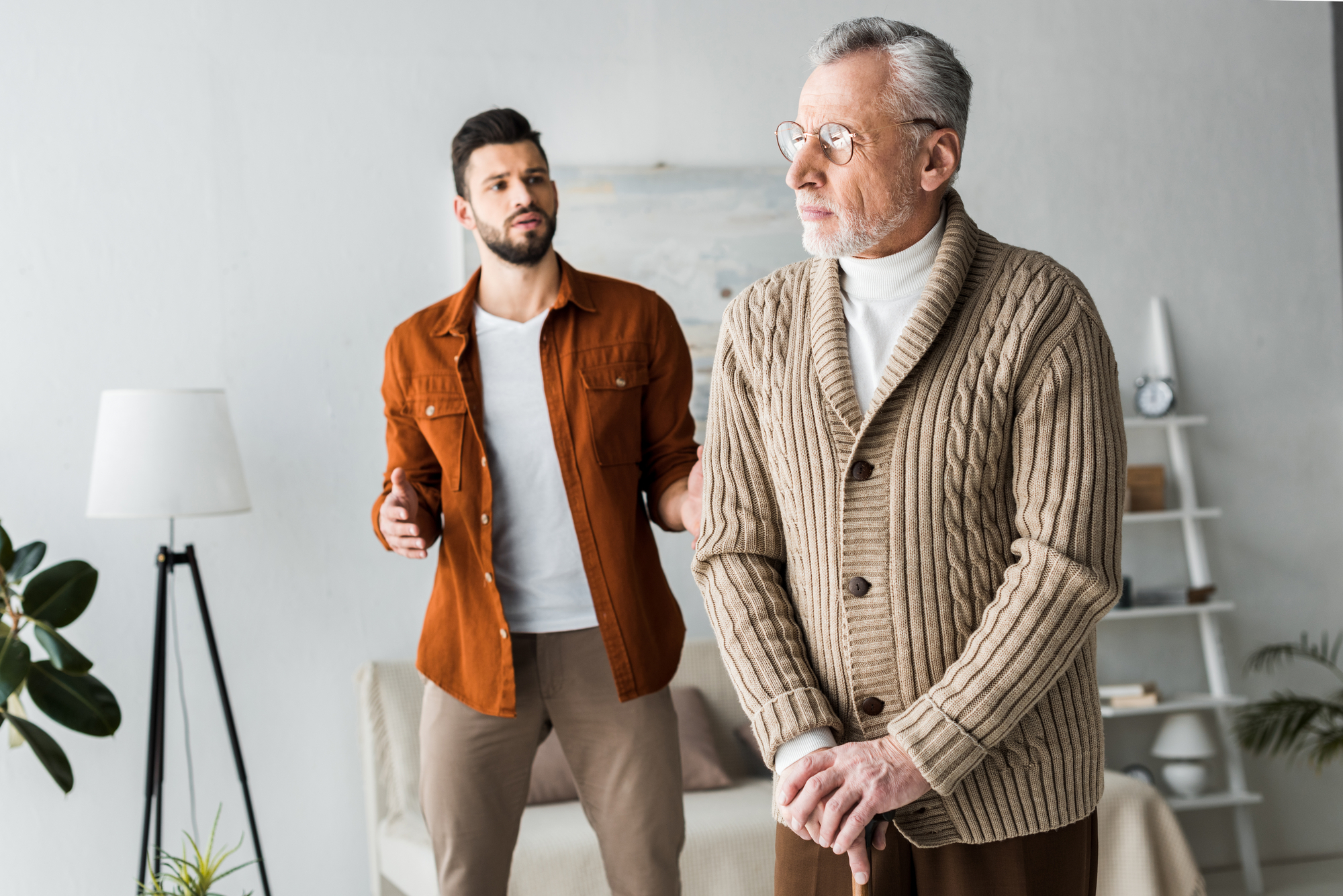 An elderly man with glasses and a beige cardigan stands in the foreground, holding a cane and looking to the side. A younger man with a beard, wearing a brown jacket, stands in the background with his hands outstretched, appearing to talk to the older man.