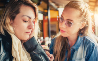 Two women sit together at a table. One woman, wearing a scarf and leather jacket, looks upset and has her head resting on her hand. The other woman, wearing glasses and a denim shirt, looks at her with a concerned expression, offering support.