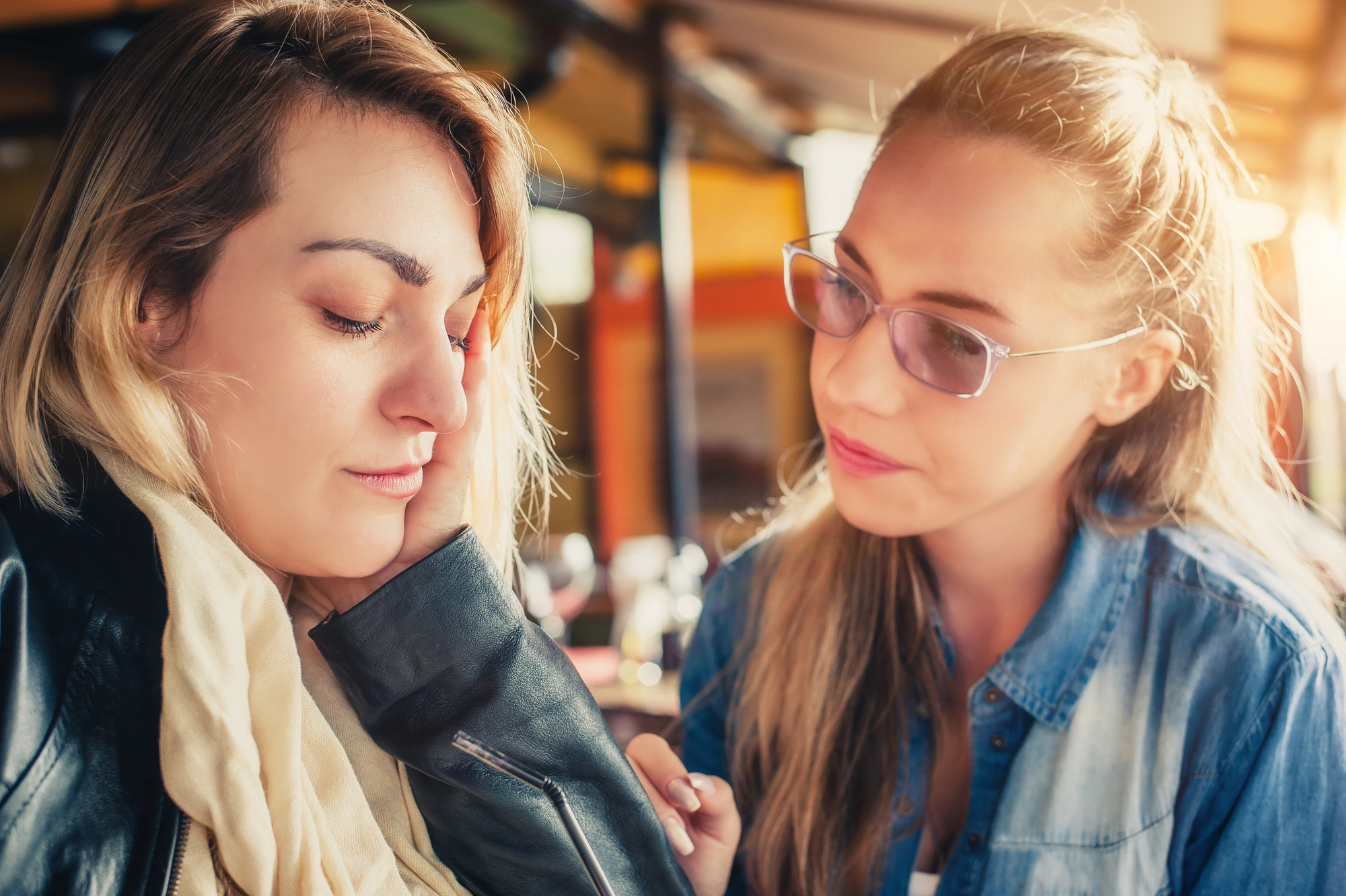 Two women sit together at a table. One woman, wearing a scarf and leather jacket, looks upset and has her head resting on her hand. The other woman, wearing glasses and a denim shirt, looks at her with a concerned expression, offering support.