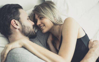 A couple lies on a bed, facing each other and smiling. The man has a beard and is wearing a gray shirt, while the woman, who has blonde hair, is in a black top. They are embracing each other lovingly.