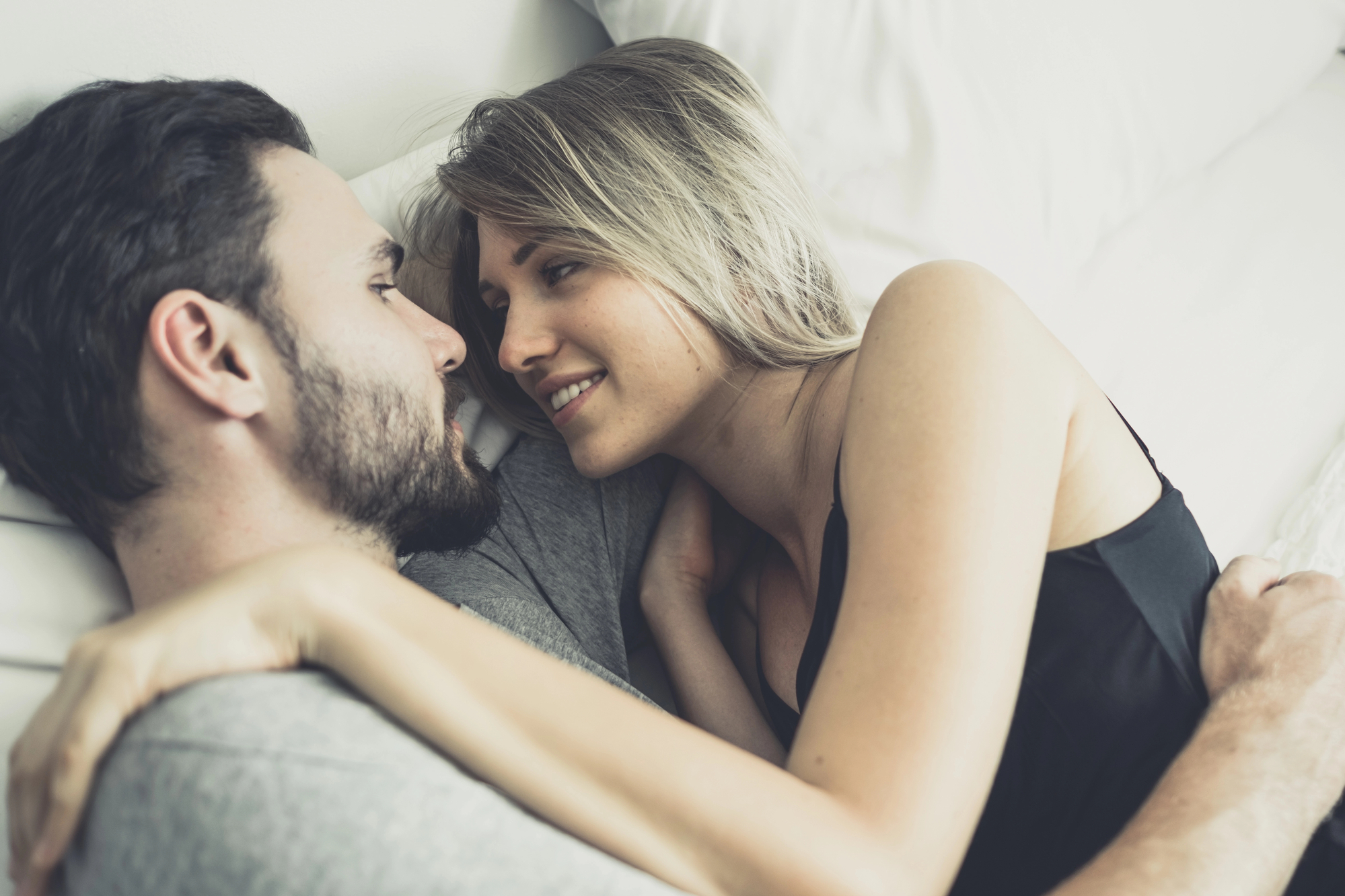 A couple lies on a bed, facing each other and smiling. The man has a beard and is wearing a gray shirt, while the woman, who has blonde hair, is in a black top. They are embracing each other lovingly.