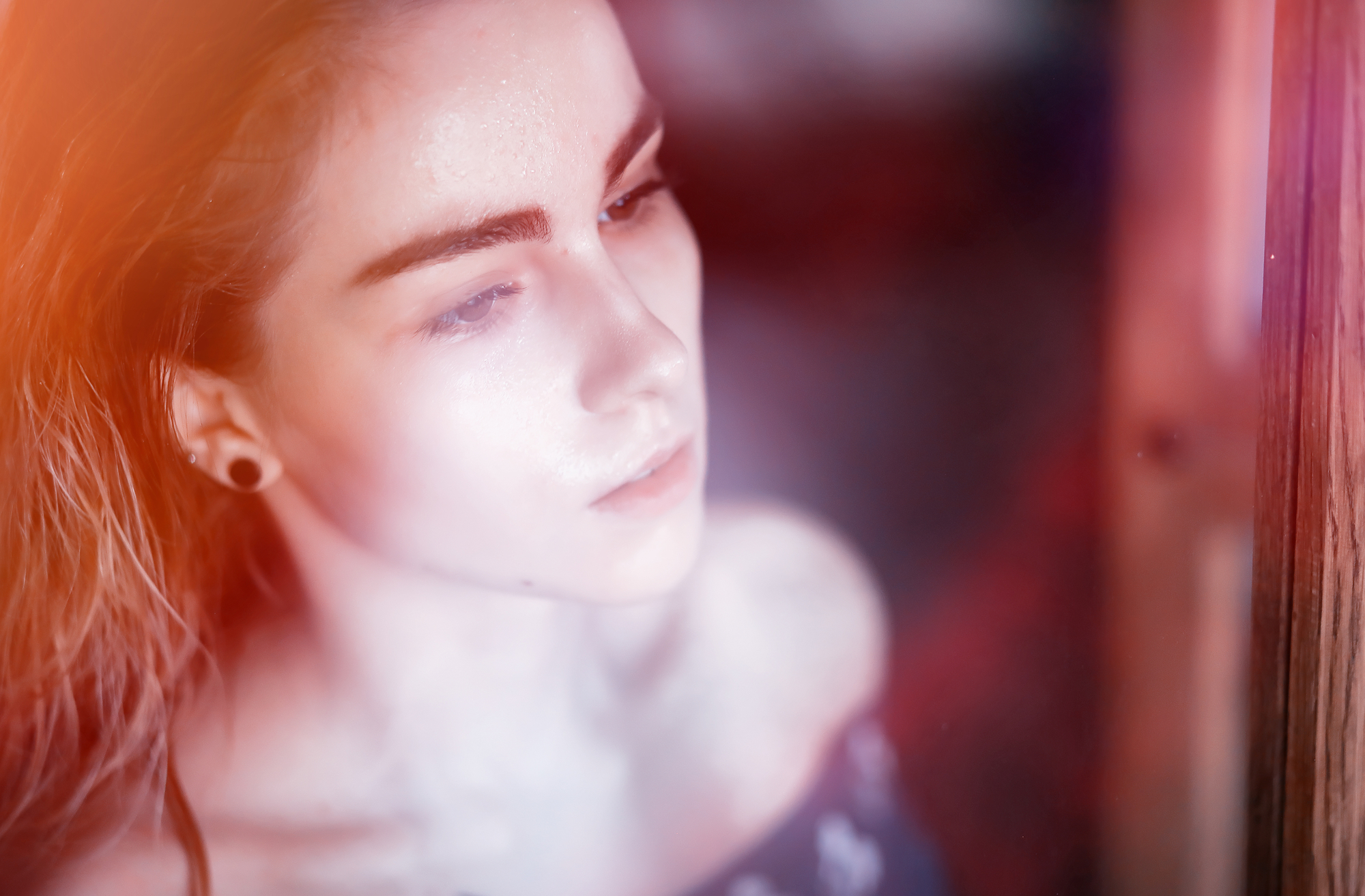 A close-up of a person with a contemplative expression, looking out a window with soft, natural light illuminating their face. The image has a dreamy, ethereal quality with a slight blur and warm tones. The person has long hair and is wearing a dark, off-the-shoulder top.