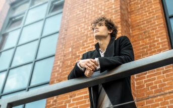 A person with curly hair wearing a black coat gazes upward while leaning on a railing. They are standing in front of a brick building with large windows. The sky is visible reflecting in the windows.