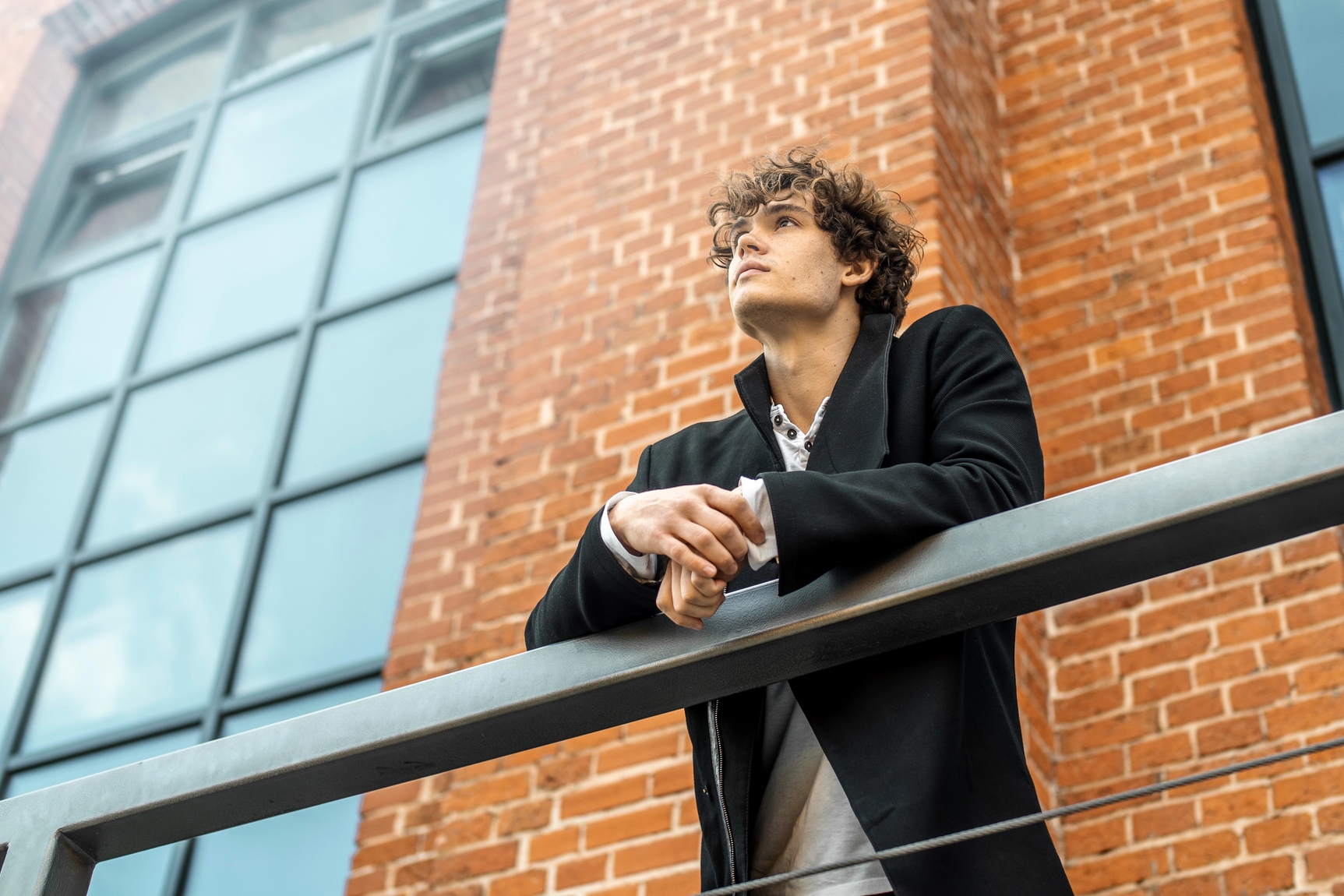 A person with curly hair wearing a black coat gazes upward while leaning on a railing. They are standing in front of a brick building with large windows. The sky is visible reflecting in the windows.