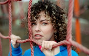 A person with curly hair and a blue hoodie stands behind a pink climbing rope net, holding the ropes with both hands and looking through the gaps in the net. The background is blurred and features outdoor play equipment in muted colors.