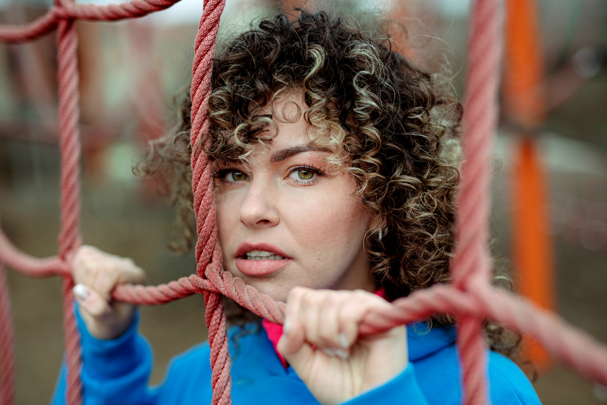 A person with curly hair and a blue hoodie stands behind a pink climbing rope net, holding the ropes with both hands and looking through the gaps in the net. The background is blurred and features outdoor play equipment in muted colors.