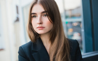 A young woman with long, straight brown hair and fair skin is looking off to the side with a neutral expression. She is wearing a dark blazer, and the background shows an urban setting with blurred buildings and vehicles.