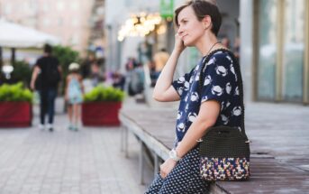 A person with short hair, wearing a patterned shirt and checkered pants, leans against a platform while holding a crocheted handbag. The street scene behind features pedestrians and a building with lights.