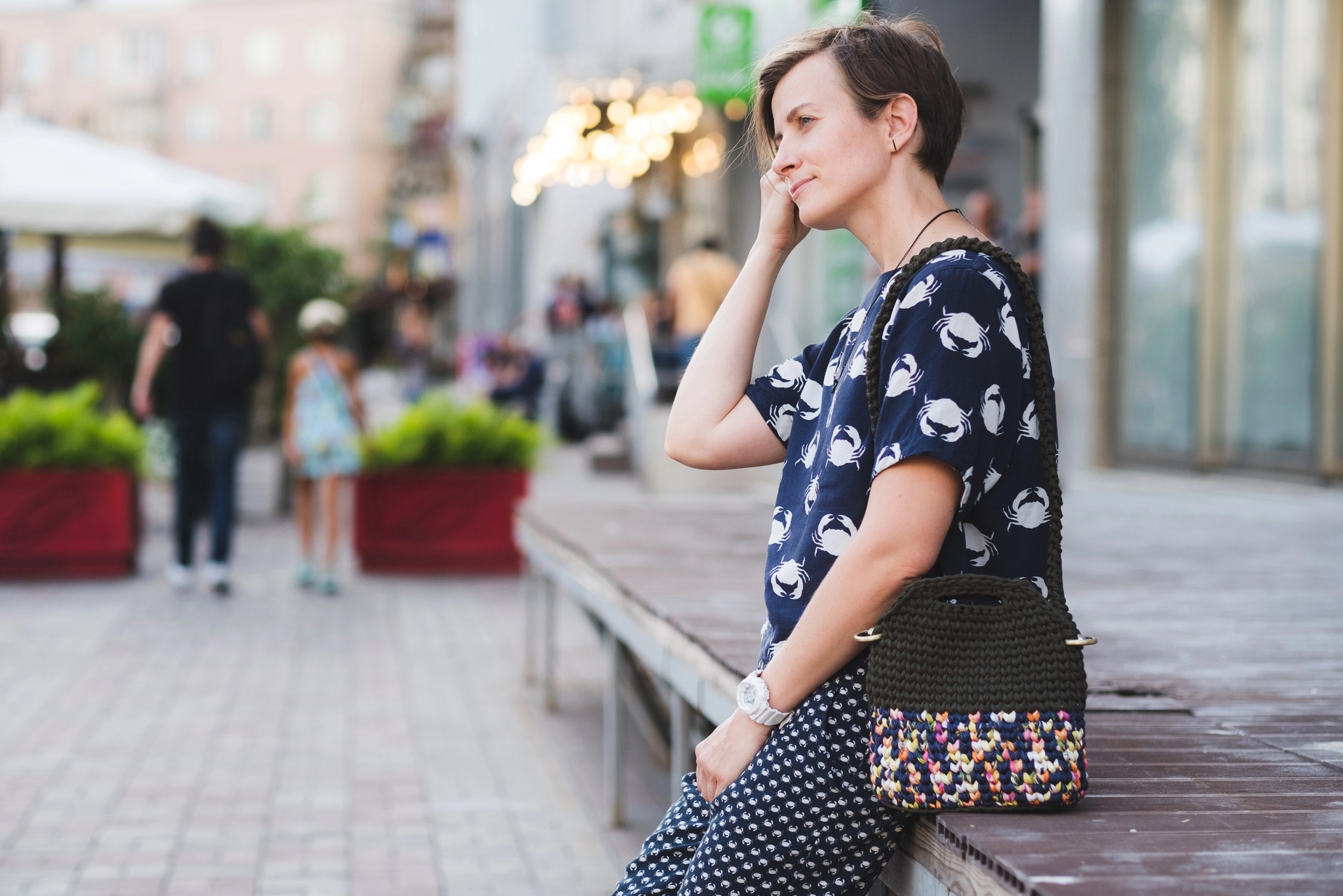 A person with short hair, wearing a patterned shirt and checkered pants, leans against a platform while holding a crocheted handbag. The street scene behind features pedestrians and a building with lights.