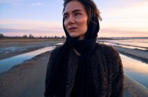 Young woman wearing dark scarf and coat, standing outdoors on a beach during sunset. The sky is painted in soft pastel colors with water and coastline in the background. She looks serene and thoughtful.