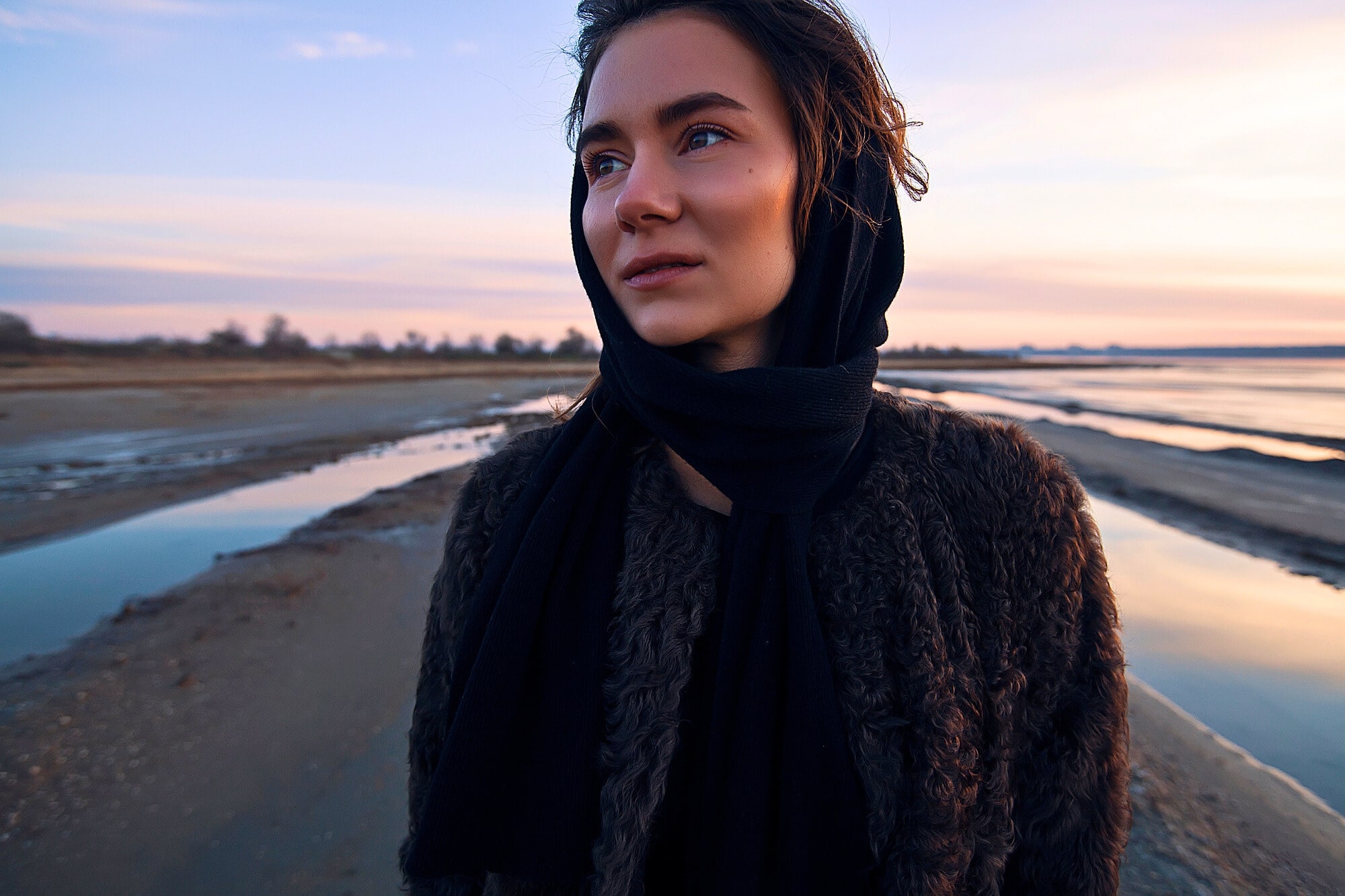 Young woman wearing dark scarf and coat, standing outdoors on a beach during sunset. The sky is painted in soft pastel colors with water and coastline in the background. She looks serene and thoughtful.