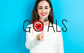 A smiling woman with long brown hair and red lipstick, wearing a white sweater, points at a target icon in the word "GOALS" on a clear screen. The background is bright blue, creating a contrasting effect with the text and her outfit.