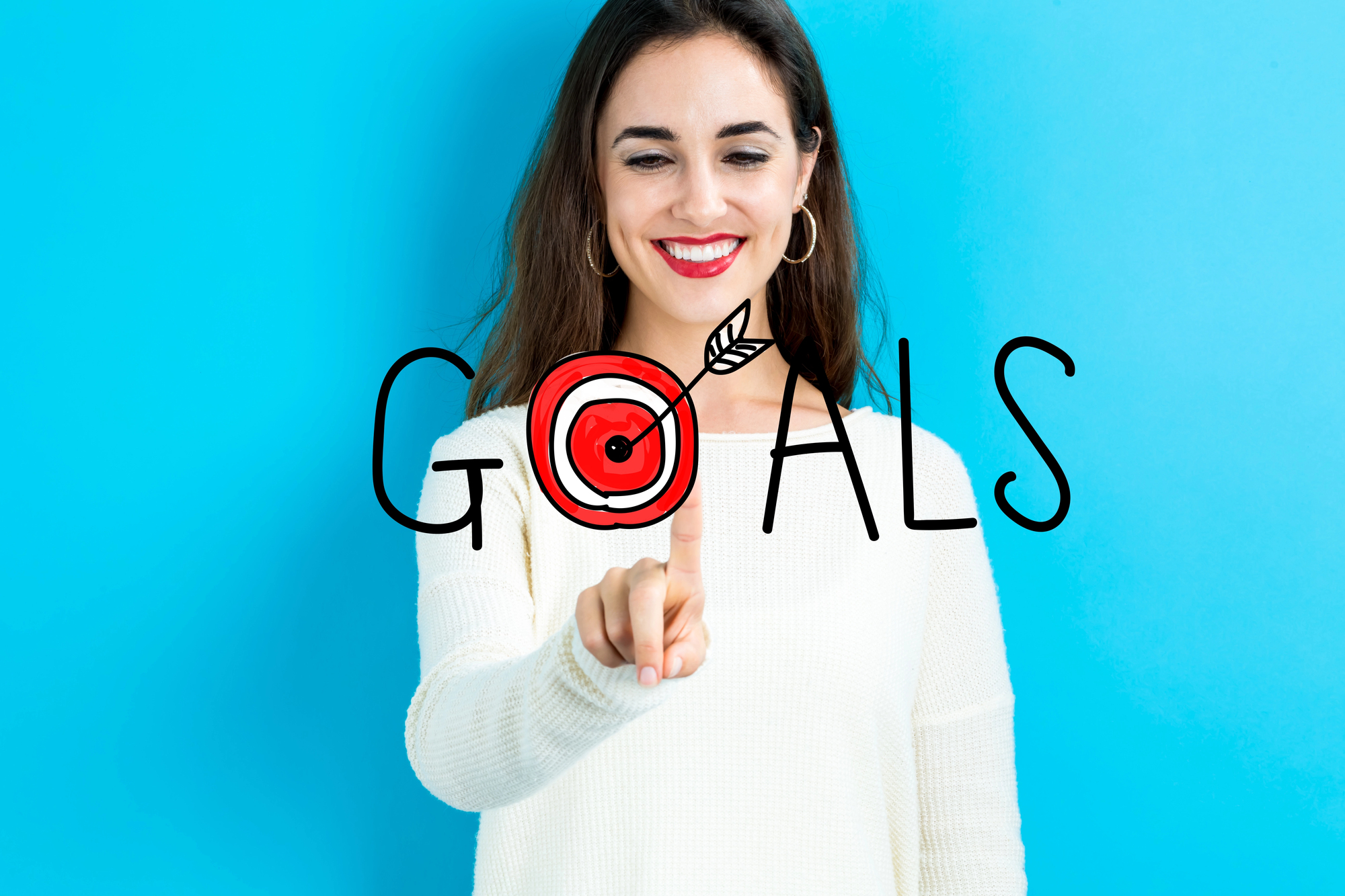 A smiling woman with long brown hair and red lipstick, wearing a white sweater, points at a target icon in the word "GOALS" on a clear screen. The background is bright blue, creating a contrasting effect with the text and her outfit.